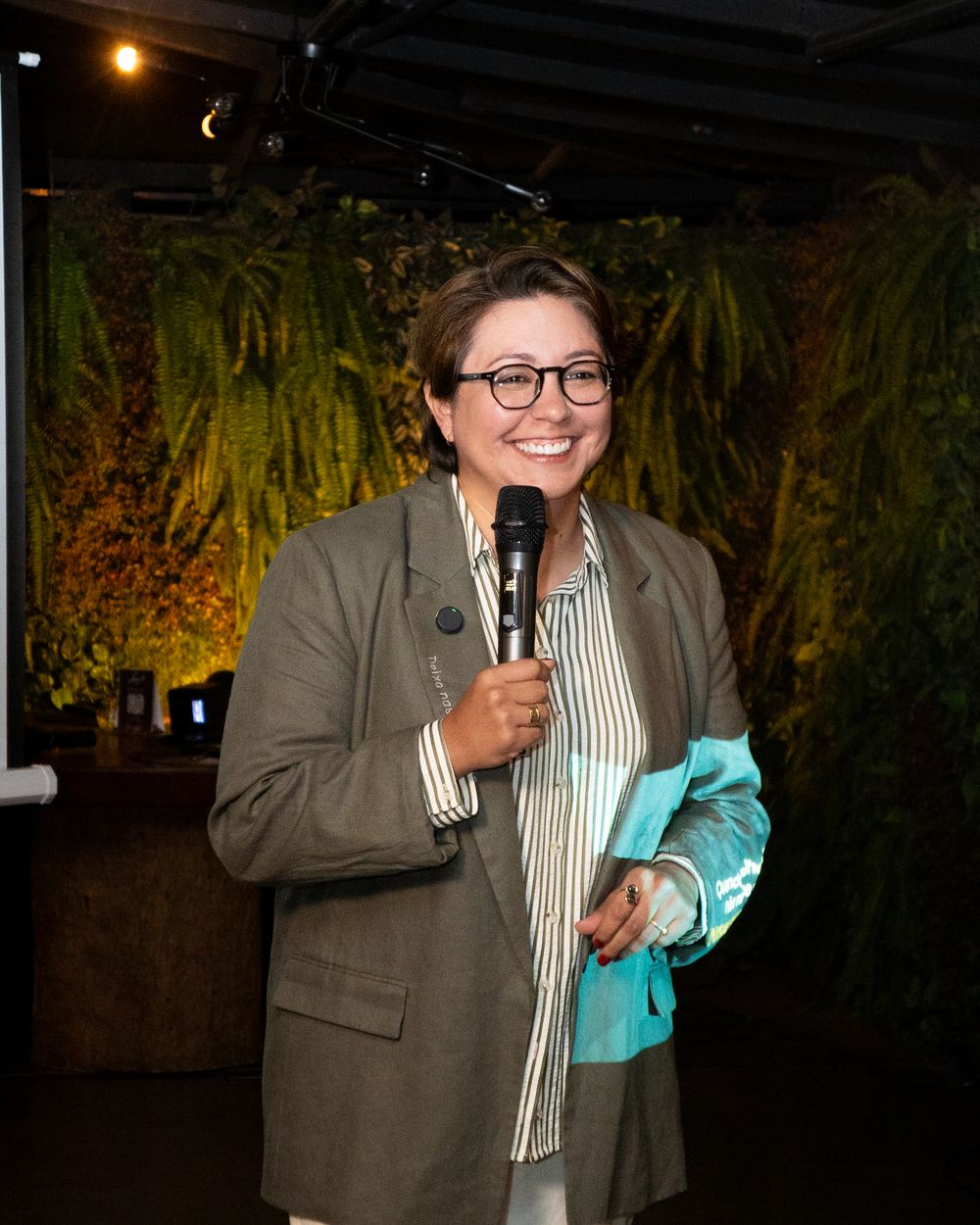 Eliesita Pereira falando ao microfone e sorrindo, com plantas ao fundo.
